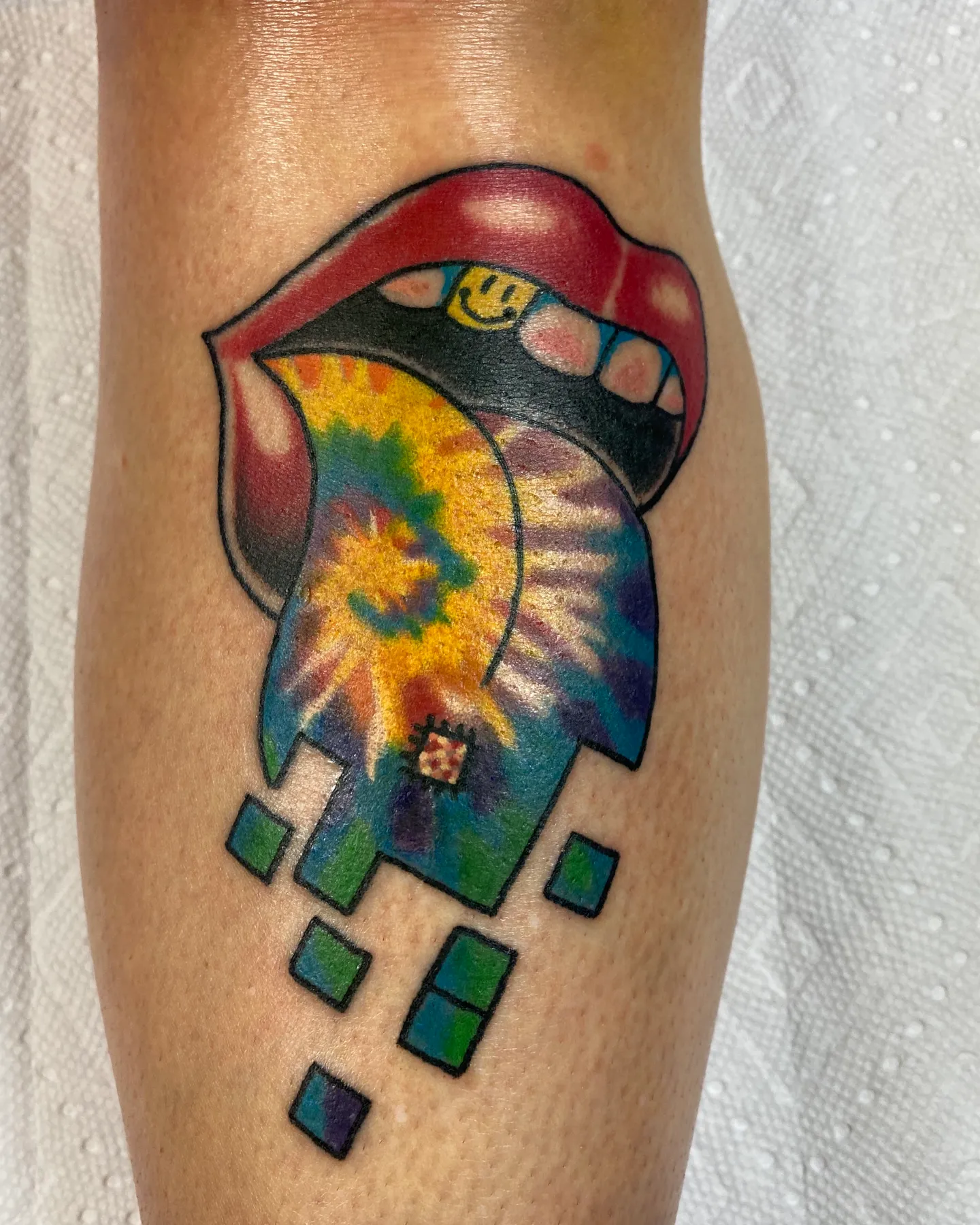 Tie-dye lips and tongue tattoo with smiley face accents by Mathias Moretti at Golden Child Tattoo in Escondido
