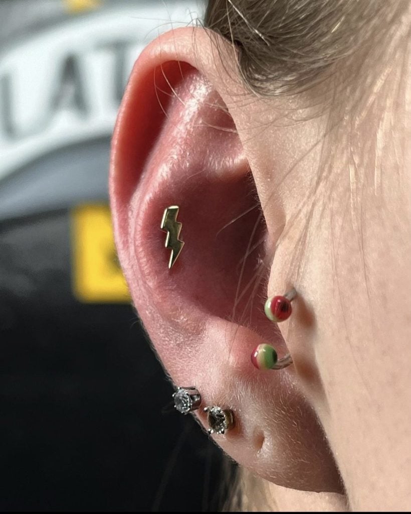 Ear piercing with a gold lightning bolt stud in the conch, crystal studs in the lobe, and colorful ball ends at the tragus, by Birdie at Golden Child Tattoo and Piercing, Escondido